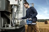A person wearing safety equipment uses a cordless grease gun to service farm machinery.