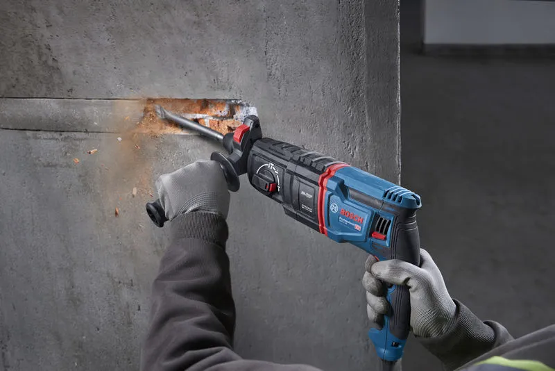 A person wearing safety equipment uses a rotary hammer to chisel into a concrete wall.