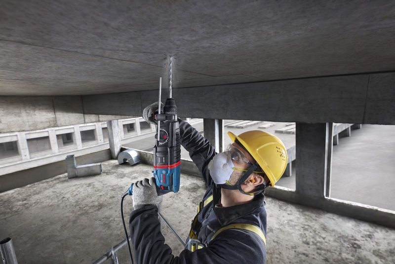 A person wearing safety equipment uses a rotary hammer to drill into a concrete ceiling.