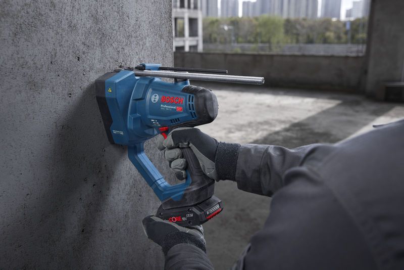 A person wearing safety equipment cuts a threaded rod against a concrete wall.