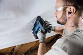 A person wearing safety equipment secures material to a ceiling using a cordless nail gun.