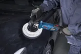 A person wearing safety equipment uses a polisher on a black car hood.
