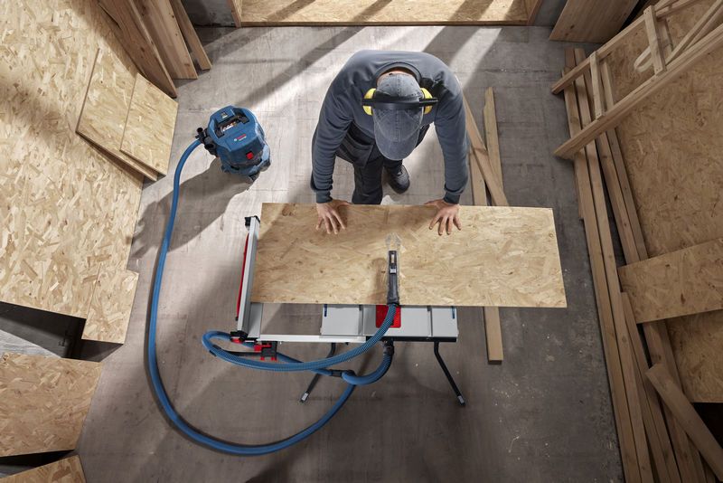 A person wearing safety equipment cuts plywood on a table saw in a workshop.