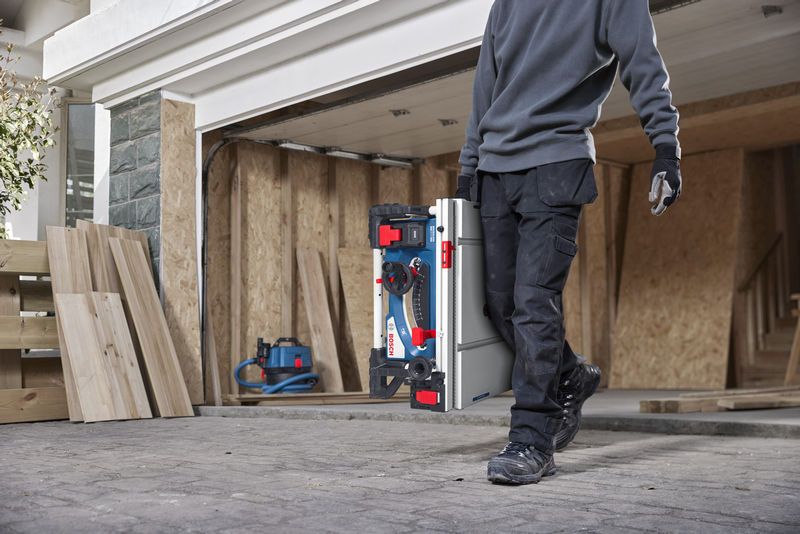 A person wearing safety equipment carries a portable table saw at a construction site.