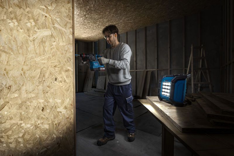 A person wearing safety equipment drills into a wooden board near a cordless jobsite light.