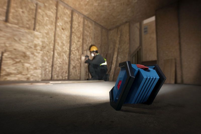 A person wearing safety equipment installs drywall, illuminated by a cordless jobsite light.