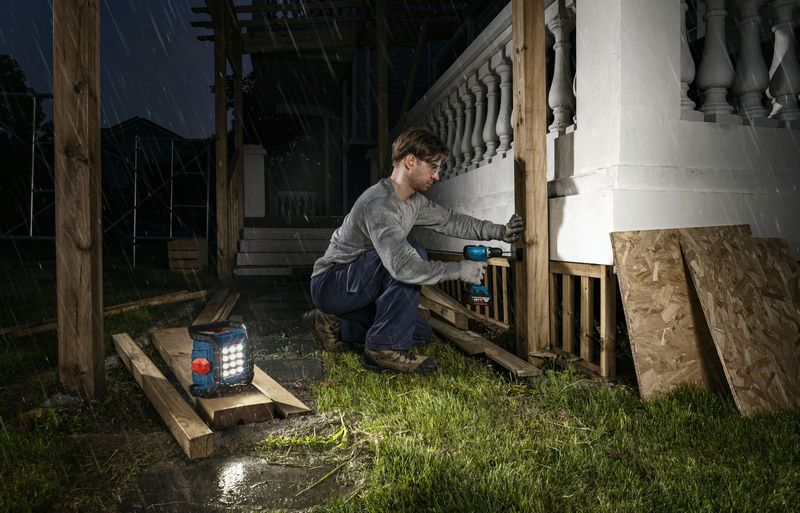 A person wearing safety equipment drills wooden posts at night with a cordless jobsite light nearby.
