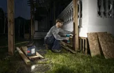 A person wearing safety equipment drills wooden posts at night with a cordless jobsite light nearby.