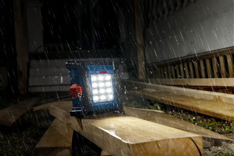 Cordless jobsite light illuminating wooden planks in heavy rain at a construction site.