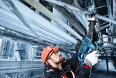 A person wearing safety equipment uses a reciprocating saw to cut a metal pipe overhead.