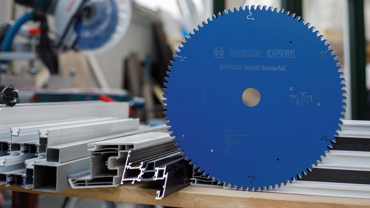 Object #200585101: 7557700_app_web_02 Large blue circular saw blade on a workbench beside stacked metal profiles.
