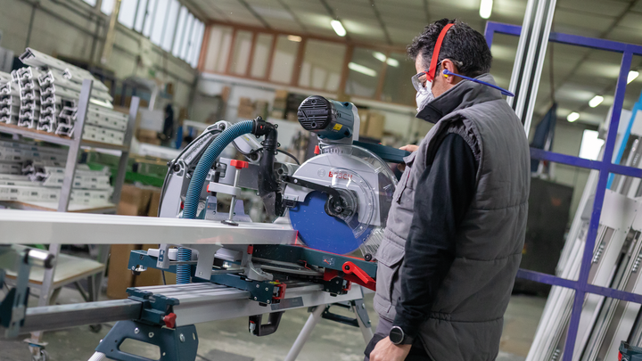 Object #200585103: 7557700_app_web_04 Person wearing safety equipment cuts metal pipe with a sliding miter saw in a workshop.