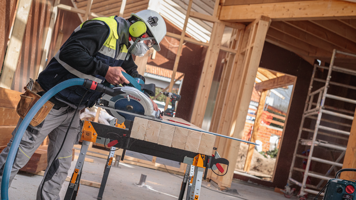 Object #200585282: 7557710_app_web_07 A person wearing safety equipment cuts wooden beams with a circular saw at a construction site.