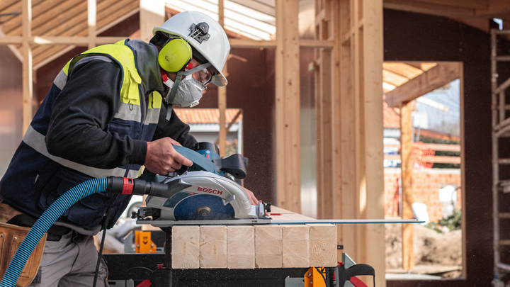 Object #200585283: 7557710_app_web_08 Person wearing safety equipment uses a circular saw to cut wood at a construction site.