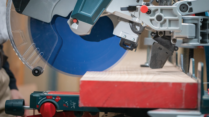 Object #200585324: 7557711_app_web_10 Miter saw with a blue blade cutting a wooden plank on a workshop table.