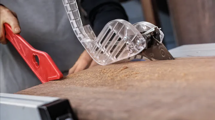 Object #200585342: 7557712_app_web_07 Person uses a push stick to guide wood through a table saw with safety guard.