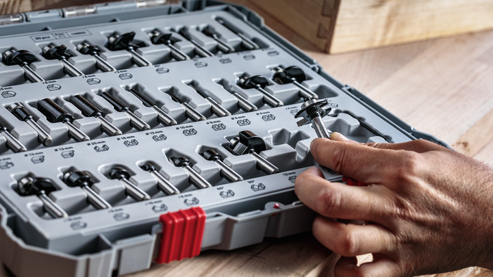 Person selecting a router bit from an organized tool case on a wooden surface.
