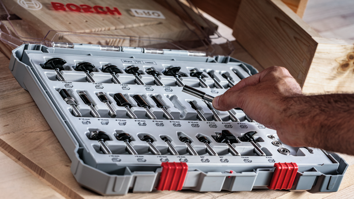 Person selecting a router bit from an organized case on a wooden table.