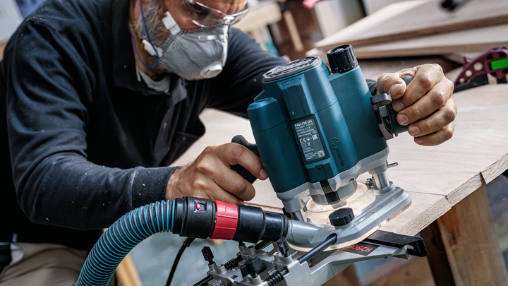 Person wearing safety equipment uses a power router to shape the edge of a wooden board.