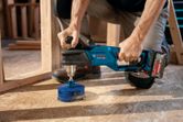 A person uses a cordless angle drill to bore a large hole in a wooden floor.