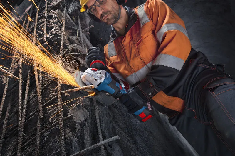 A worker wearing safety equipment uses an angle grinder to cut metal rebar.