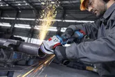 A person wearing safety equipment grinds a metal pipe with an angle grinder, creating sparks.