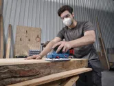 A person wearing safety equipment sands a wooden board with a cordless orbital sander.