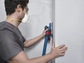 A person wearing safety equipment sands a wall with an orbital sander connected to a vacuum hose.