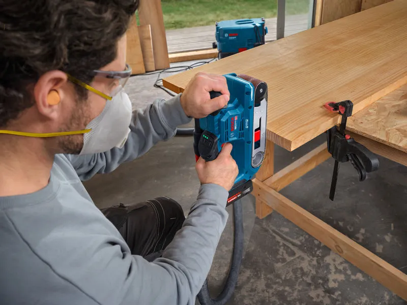 A person wearing safety equipment sands a wooden board using a belt sander.