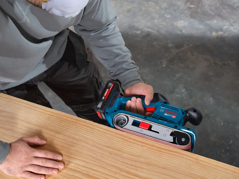 A person wearing safety equipment sands a wooden board with a cordless belt sander.