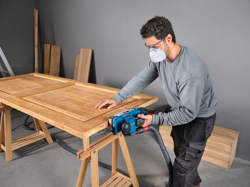 A person wearing safety equipment sands a wooden door with a belt sander.