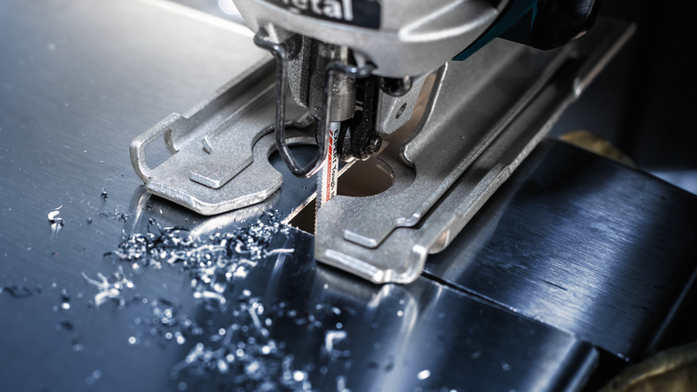 EXPERT Tough Metal thin T118AHM Jigsaw Blade, 3 pcs Jigsaw cutting a sheet of metal, small metal shavings scattered around the blade.