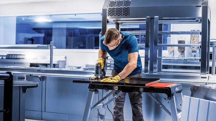 Object #200597847: 2793293_app_web_05 A person wearing safety equipment grinds a metal sheet on a workbench in a kitchen.