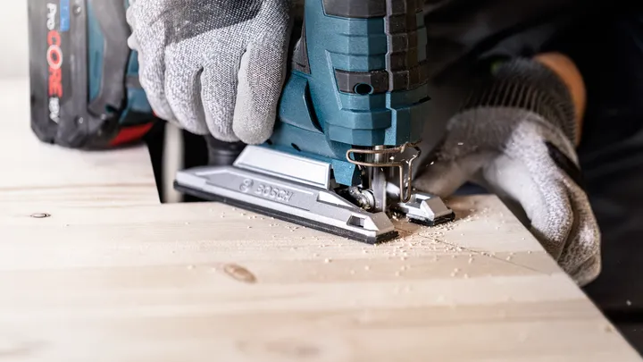 Person wearing safety equipment uses a cordless jigsaw to cut a wooden board.