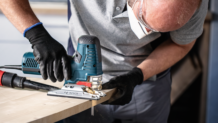 Person wearing safety equipment cuts wood using a power jigsaw.