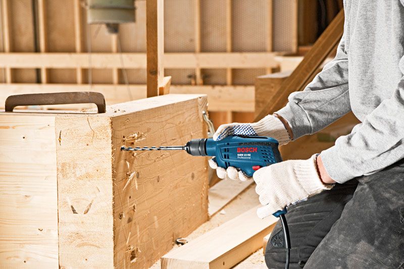A person wearing safety equipment drills into a wooden beam using a power drill.