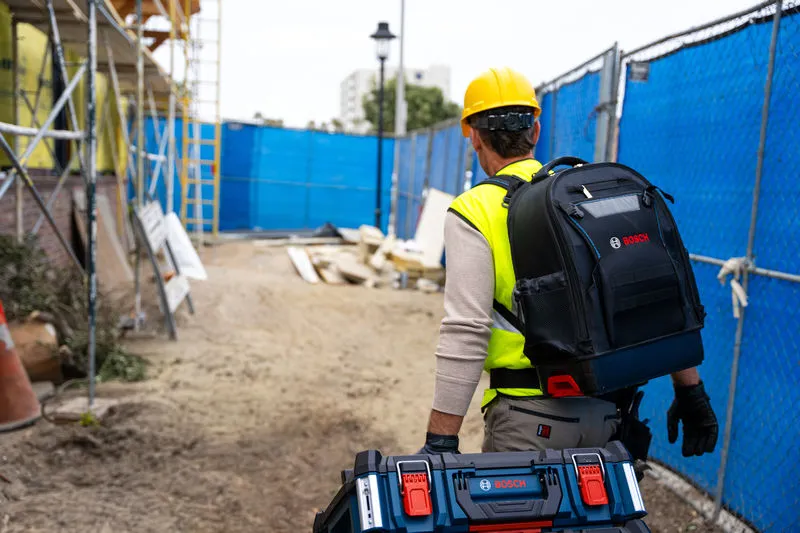 A person wearing safety equipment carries a contractor backpack and carrying case system at a construction site.