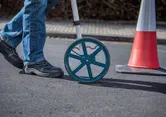 A person wearing safety equipment uses a measuring wheel on a paved surface.