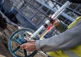 A person wearing safety equipment measures distance with a large measuring wheel.