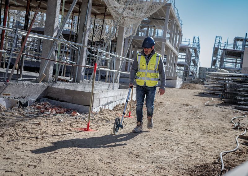 A person wearing safety equipment uses a measuring wheel at a construction site.