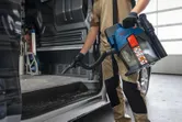 A person wearing safety equipment cleans a vehicle interior using a cordless dust extractor.