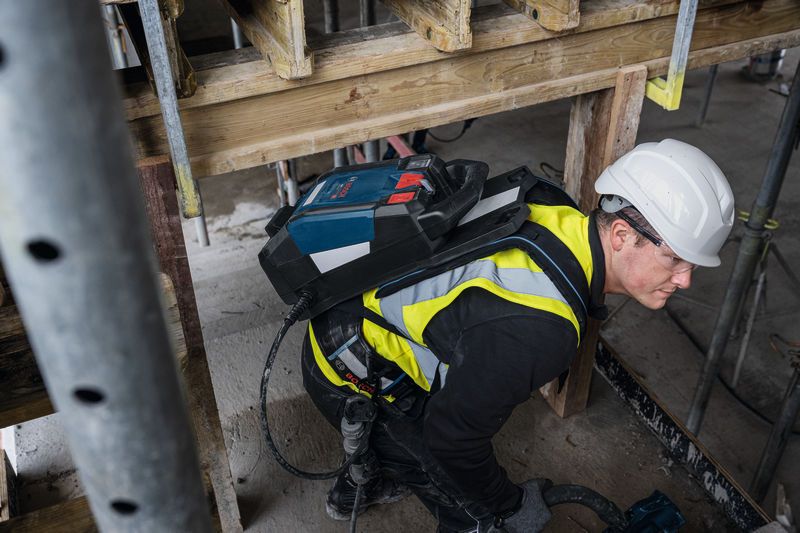 A person wearing safety equipment uses a tool while carrying a power supply backpack.