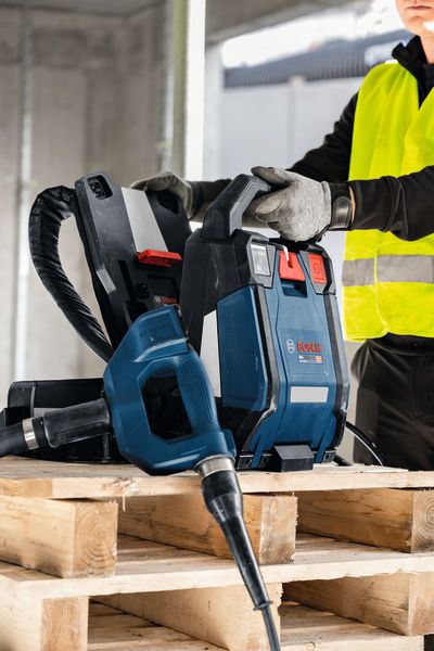 A person wearing safety equipment operates a dust extractor and drill on wooden pallets.