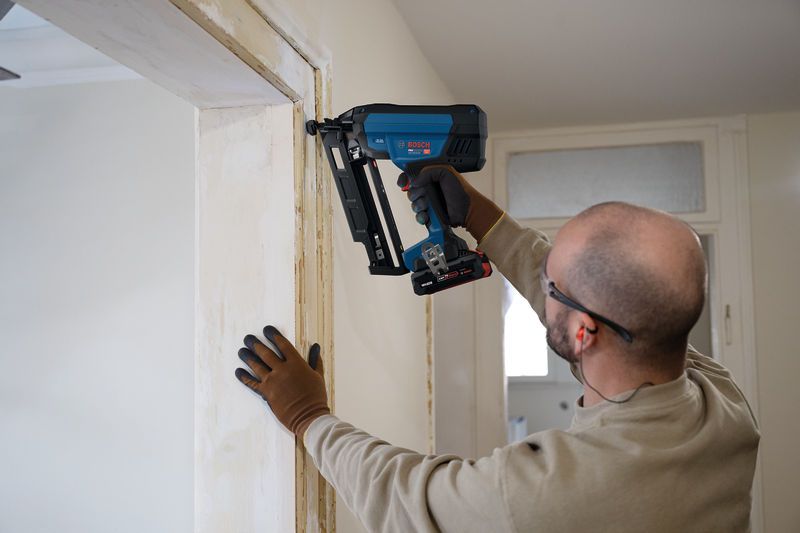 A person wearing safety equipment uses a cordless finish nailer on a door frame.