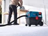 A person wearing safety equipment uses a dust extractor while cutting wood on a table.