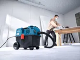 A person wearing safety equipment uses an all-purpose extractor while cutting wood on a table.