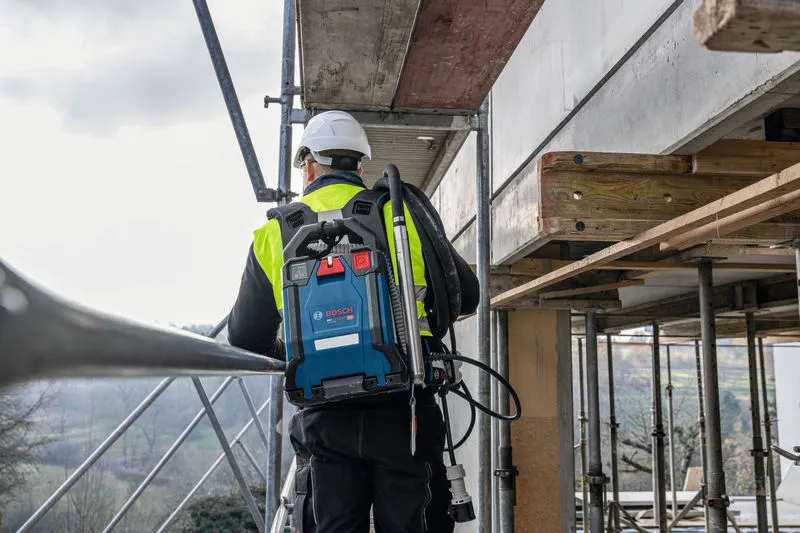 Person wearing safety equipment stands on scaffolding outdoors with a blue backpack vacuum system.