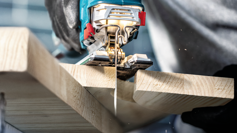 Person wearing safety equipment cuts a plank of wood with a jigsaw.