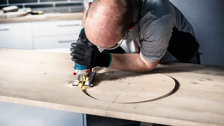 Person wearing safety equipment cuts a circular hole in wood using a jigsaw.