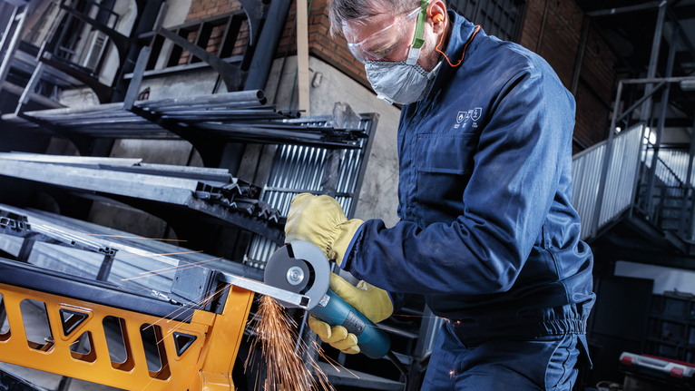 Person wearing safety equipment uses an angle grinder to cut a metal beam.